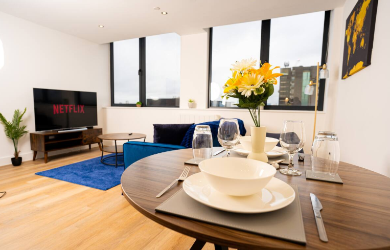 Bright modern apartment with blue sofa, wooden dining table set with wine glasses, Netflix on TV, and large windows with natural light