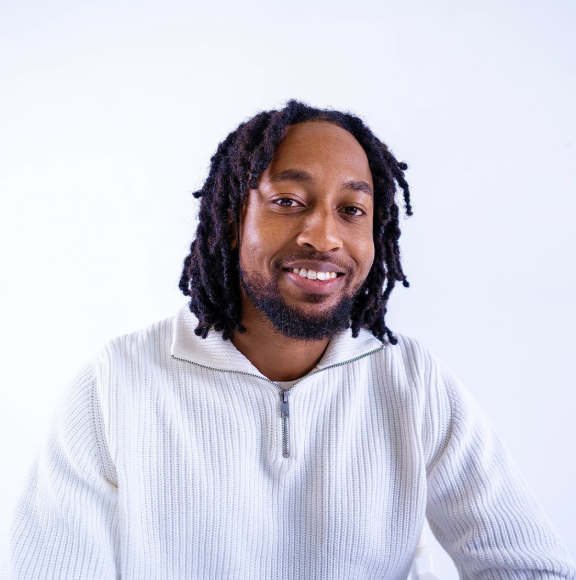 Professional portrait of Joel in white ribbed zip-up hoodie with dreadlocks, smiling warmly against light background
