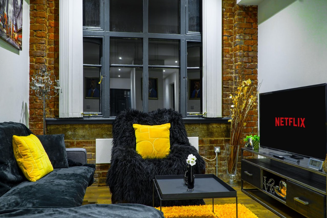 Industrial-style living room with exposed brick walls, dark velvet sofas with yellow cushions, black fluffy armchair, and Netflix on large TV